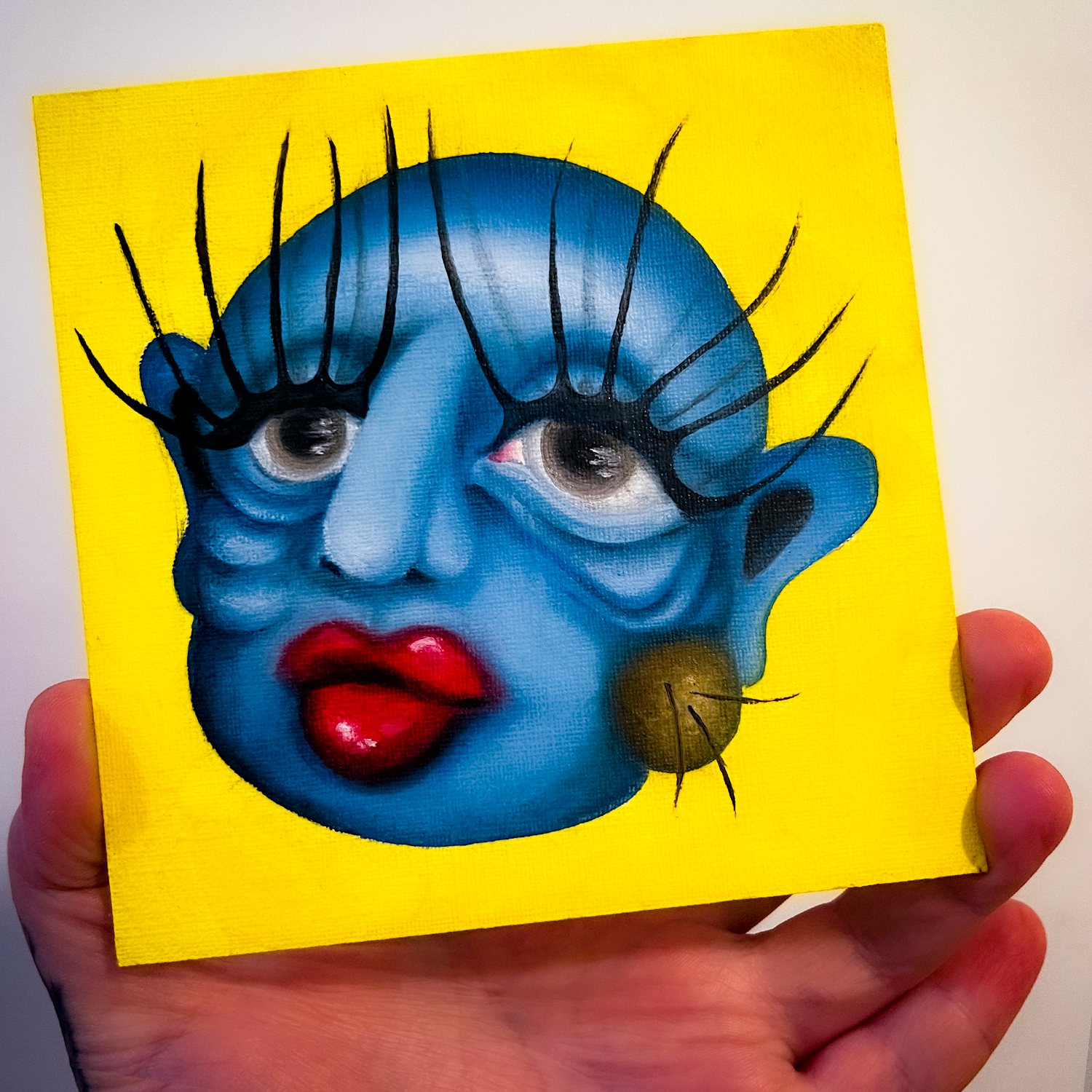 A hand holding a small painting of a face of a blue-skinned person with sagging eye bags, a large mole on the right cheek, large glossy red lips, and large eyelashes that extend upward far above their head. The background is neon yellow
